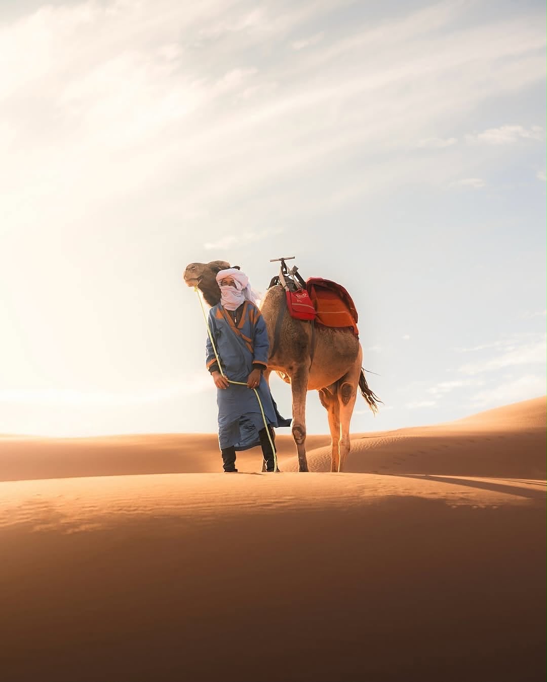 Camel Ride Camel guide in a blue turban walking with a camel on the dunes – Merzouga